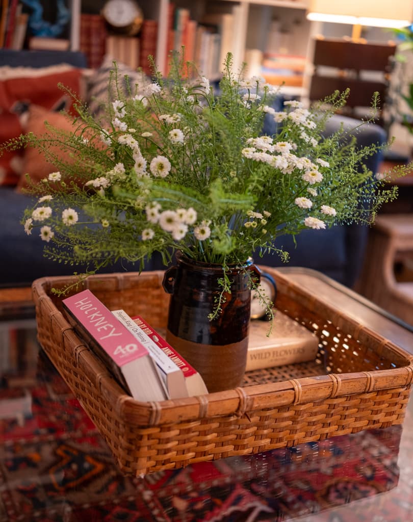 A jug of white wildflowers and a small stack of books in a woven tray on a living room table.