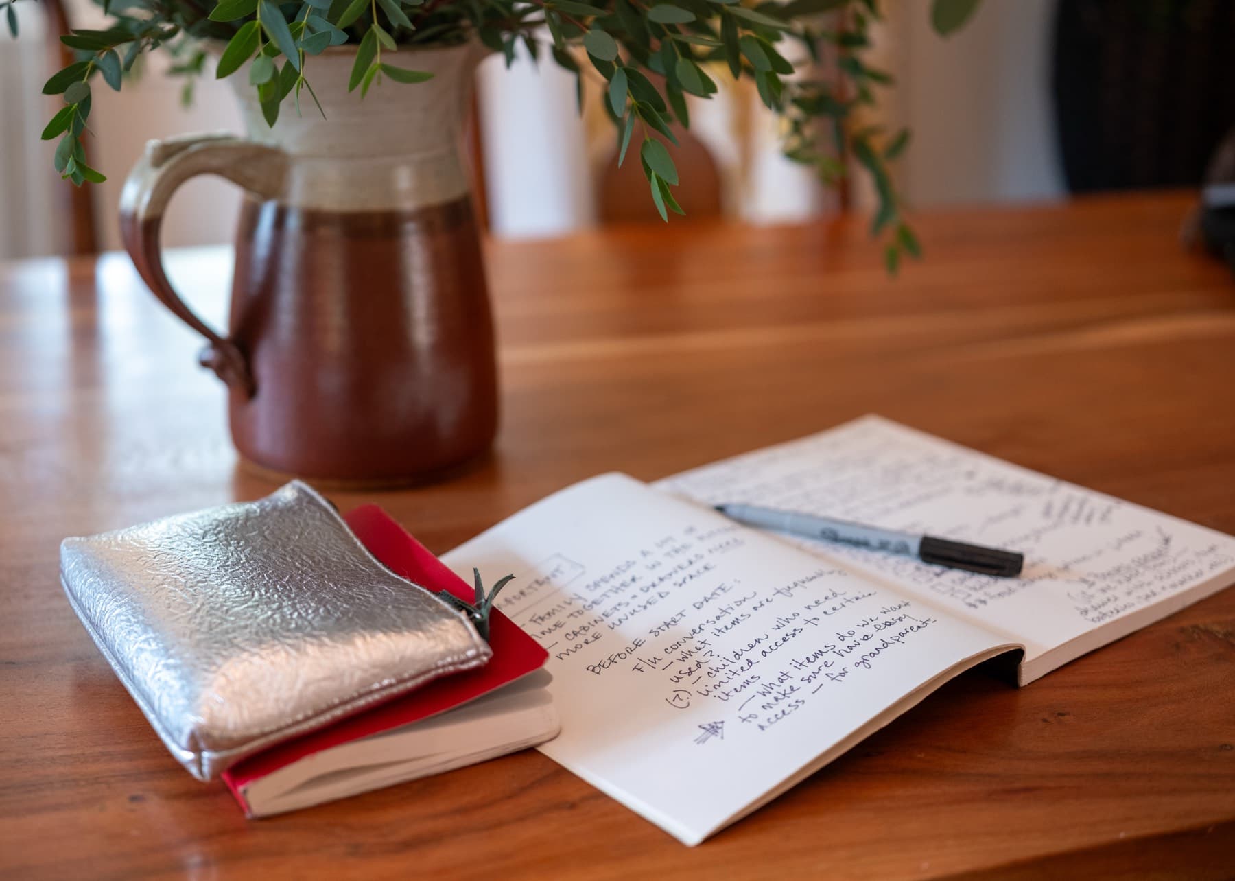An open notebook of handwritten project-planning notes with a marker resting on it, beside a pitcher of eucalyptus on a wooden table.
