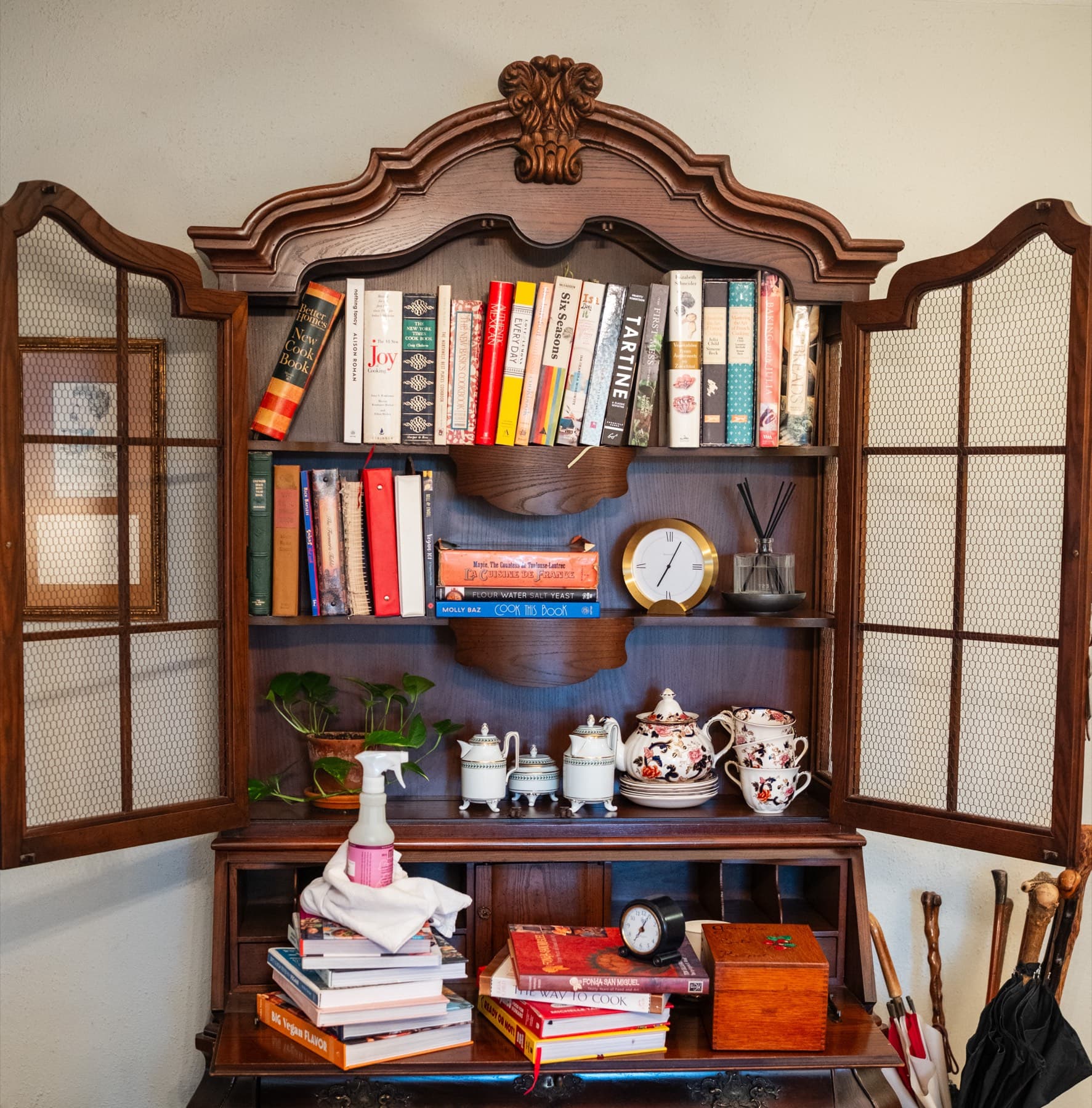 An antique glass-front cabinet with cookbooks lined up on its shelves, a teapot and cups below, and more books stacked on the desk beneath.