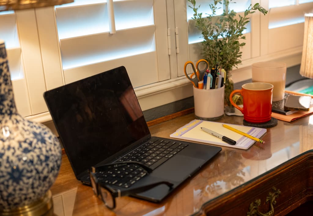 A laptop, notepad and coffee mug on a polished wooden desk beside a window.